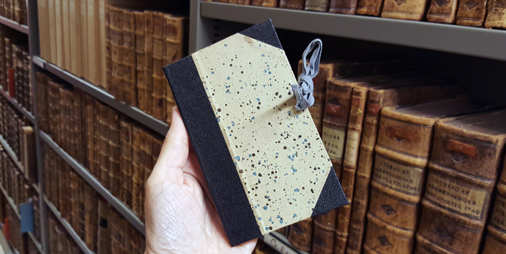 en hand håller en kaspel i form av en bok bunden i prickigt bokpapper och med ett knytsnöre över främre snittet