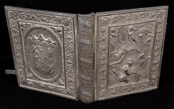 en uppslagen pärm av silver med en mängd dekor i relief, på framsidan en naken dam och på baksidan en vapenskäld